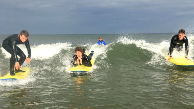 Schüler der Royal School Cavan beim Surfen auf Wellen mit Meer und bewölktem Himmel im Hintergrund.