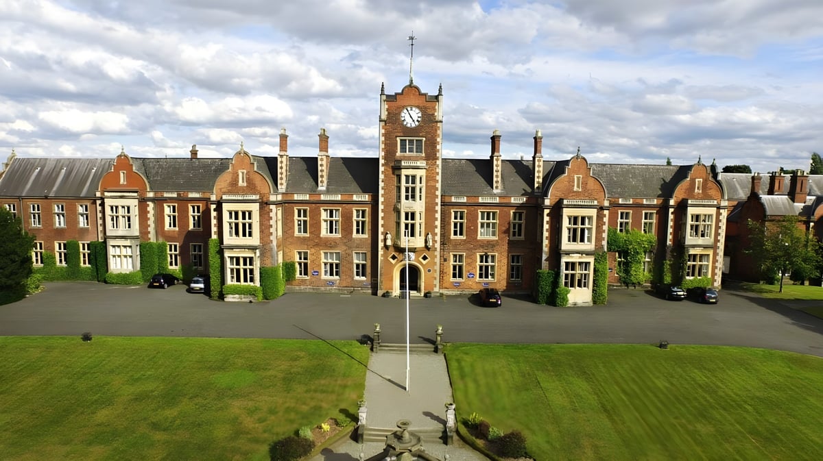 Das historische Gebäude mit Uhrenturm steht im grünen Innenhof der Royal School unter bewölktem Himmel.