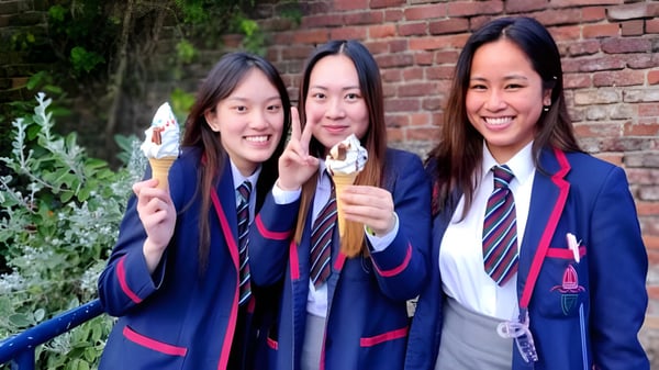 Drei Schülerinnen der Royal School tragen Schuluniformen und halten Eis in einem Garten mit Backsteinmauer.