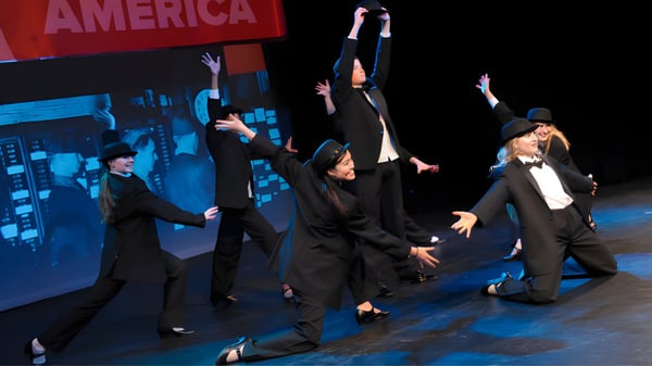 Eine Gruppe von Schülern der Rugby School tanzt und gestikuliert auf einer Bühne mit dem Schriftzug America im Hintergrund.