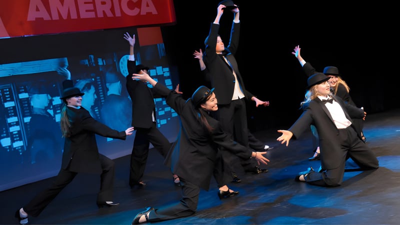 Eine Gruppe von Darstellerinnen und Darstellern der Rugby School tanzt auf der Bühne vor einem großen America-Schriftzug.