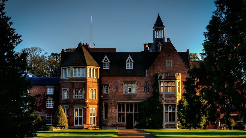 Das historische Backsteingebäude der Rugby School ist von Rasen und Bäumen umgeben und steht vor klarem blauem Himmel.