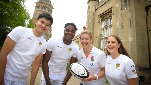 Eine Gruppe von vier Schülern in weißen Sporttrikots steht vor einem historischen Gebäude mit hohem Turm auf dem Campus der Rugby School.