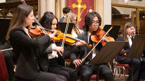 Eine Gruppe Musiker spielt verschiedene Streichinstrumente auf der Bühne der Ryde School vor rotem und goldenem Hintergrund.