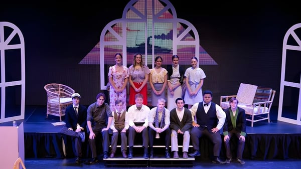 Schülerinnen und Schüler des Sacred Heart College stehen in formeller Kleidung vor einer bunten Bühne mit Bogenfenstern und architektonischen Details.