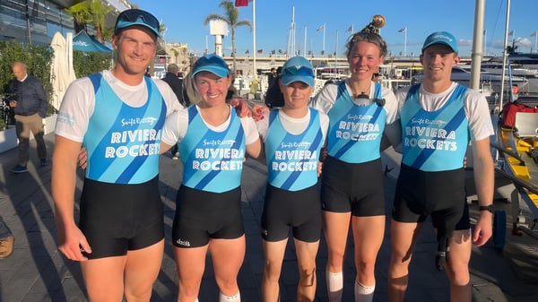 Eine Gruppe von Schülern des Sacred Heart College steht in blauen und weißen Uniformen vor einem Hafen mit Booten und Gebäuden.