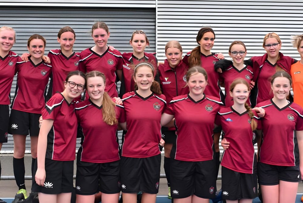 Eine Gruppe Schülerinnen des Sacred Heart College Napier posiert in roten und schwarzen Sportuniformen vor einer Metalljalousie.