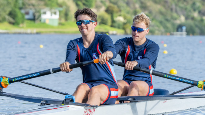 Zwei Schülerinnen in sportlichen Uniformen sitzen in einem Ruderboot auf dem Wasser vor einem bewaldeten Ufer am Sacred Heart College.
