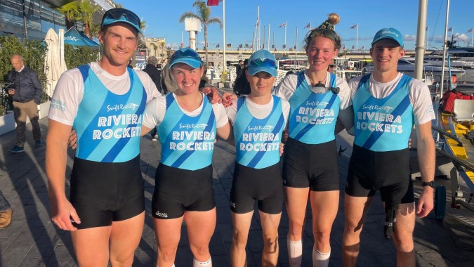 Eine Gruppe von Schülerinnen und Schülern des Sacred Heart College steht in blauen und weißen Sportuniformen vor einem Yachthafen mit Booten und Flaggen.