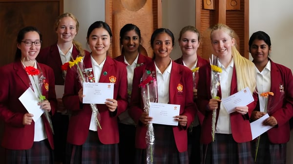 Schülerinnen des Sacred Heart Girls' College Hamilton tragen rote Abschlussthrunen und halten Zertifikate in einem Raum mit Holzpaneelen.