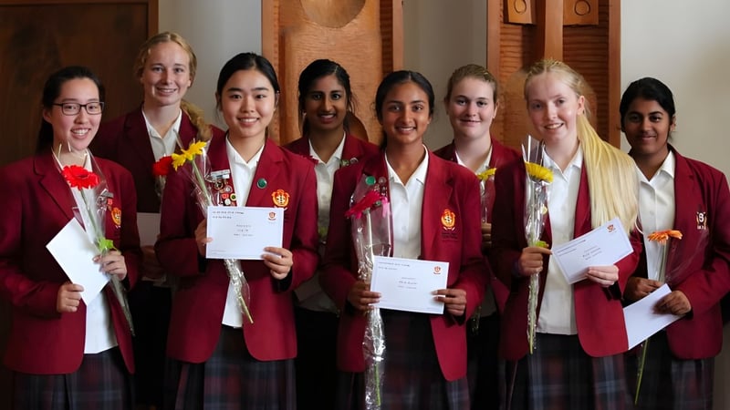 Schülerinnen des Sacred Heart Girls' College Hamilton tragen rote Abschlussthrunen und halten Zertifikate in einem Raum mit Holzpaneelen.