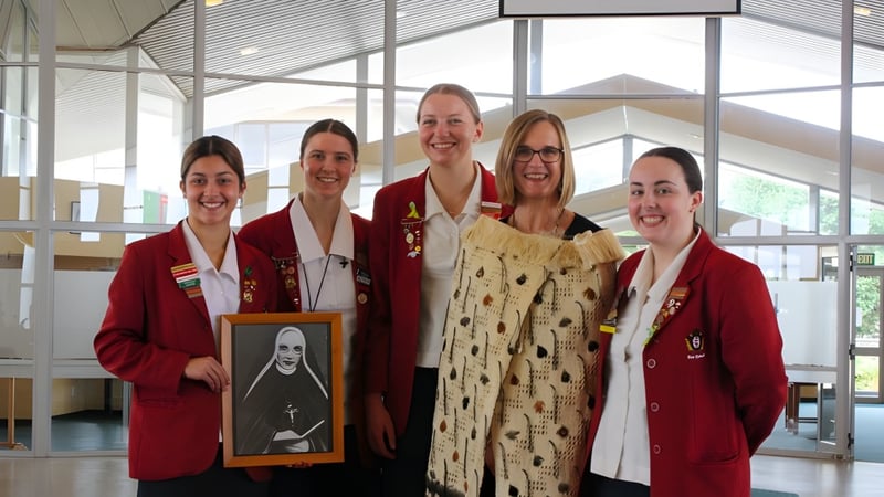 Fünf Schülerinnen in roten Talaren im hellen, verglasten Raum auf dem Campus des Sacred Heart Girls' College in New Plymouth, eine hält ein gerahmtes Porträt.