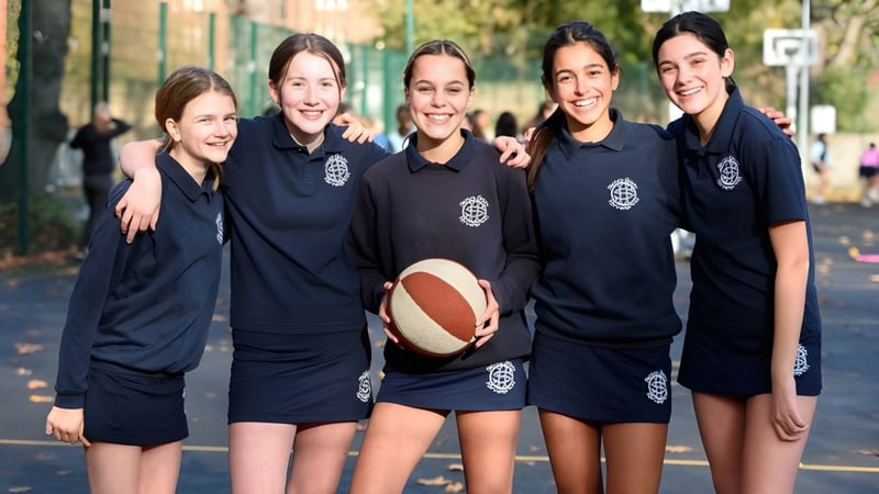 Eine Gruppe Schülerinnen der Sacred Heart High School steht mit einem Basketball in einem parkähnlichen Bereich mit Bäumen und Gebäuden.
