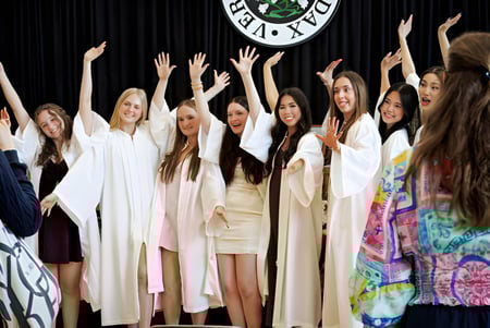 Eine Gruppe junger Schülerinnen der Sacred Heart School of Halifax steht in weißen Gewändern auf einer Bühne mit erhobenen Armen vor einem großen Logo.