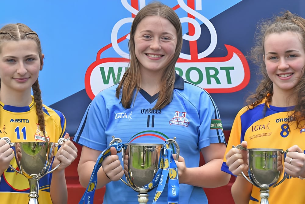 Drei Schülerinnen der Sacred Heart School in Sportuniformen halten vor einem Waterford-Logo Pokale.