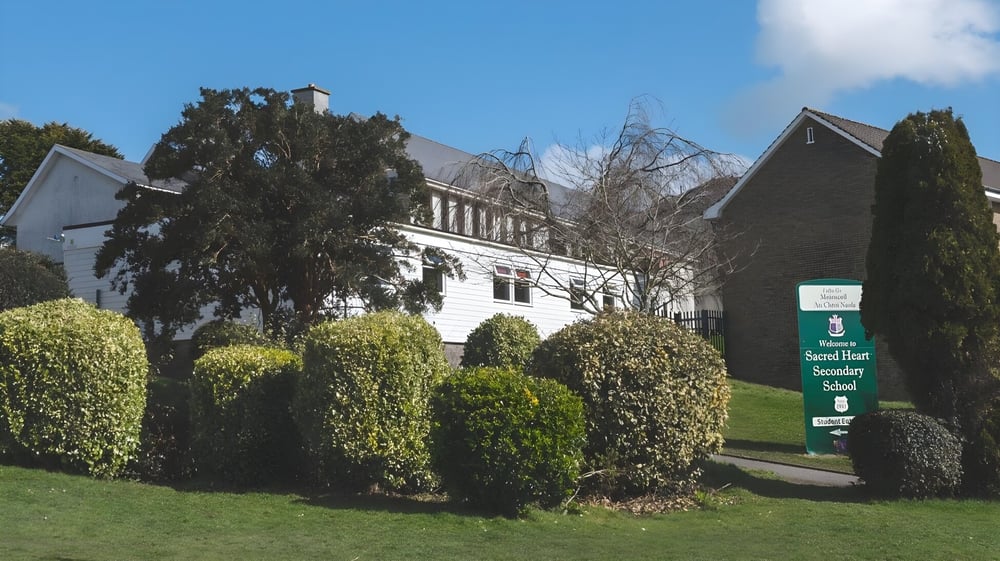 Das historische Gebäude der Sacred Heart Secondary School Clonakilty umgeben von gepflegten Hecken und Bäumen unter einem klaren blauen Himmel.