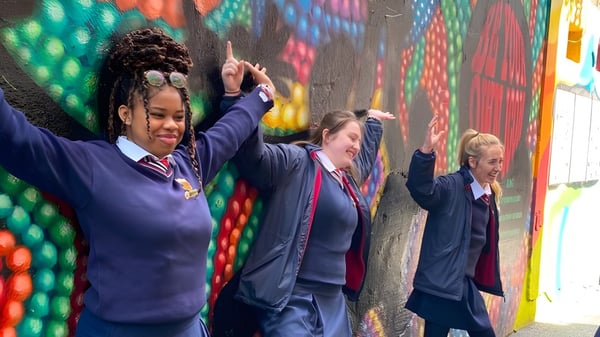 Eine Gruppe Schülerinnen und Schüler der Sacred Heart Secondary School in Schuluniform steht vor einem bunten Wandbild und hebt die Arme.