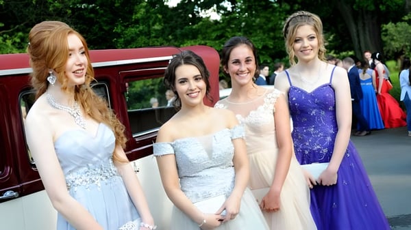 Vier Schülerinnen der Sacred Heart Secondary School Clonakilty stehen in formellen Kleidern vor einem roten Oldtimer unter Bäumen.