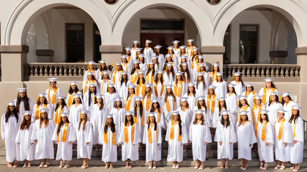Schülerinnen und Schüler der Sacred Hearts Academy stehen in weißen Roben mit gelben Schärpen vor einem verzierten Gebäude mit Säulen.