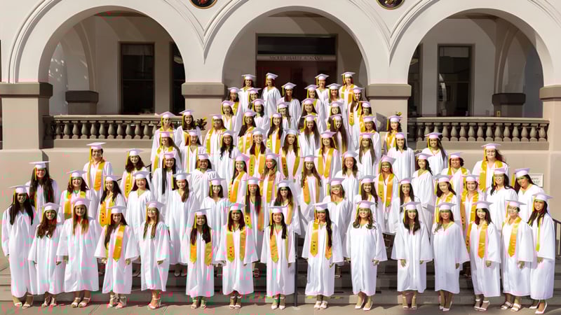Schülerinnen und Schüler der Sacred Hearts Academy stehen in weißen Roben mit gelben Schärpen vor einem verzierten Gebäude mit Säulen.