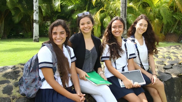 Vier Schülerinnen der Sacred Hearts Academy lächeln und posieren gemeinsam in einem tropischen Garten mit Palmen.