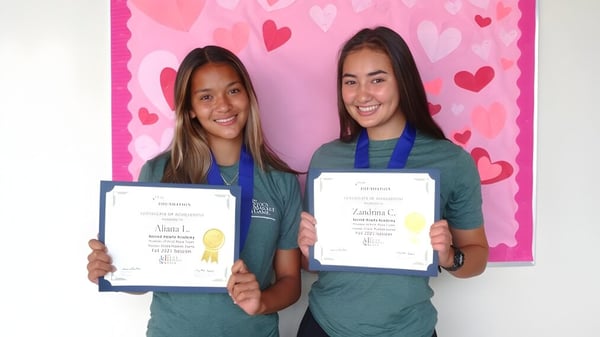 Zwei Schülerinnen der Sacred Hearts Academy halten vor einem rosafarbenen Hintergrund mit Herzmotiven ihre Zertifikate hoch.