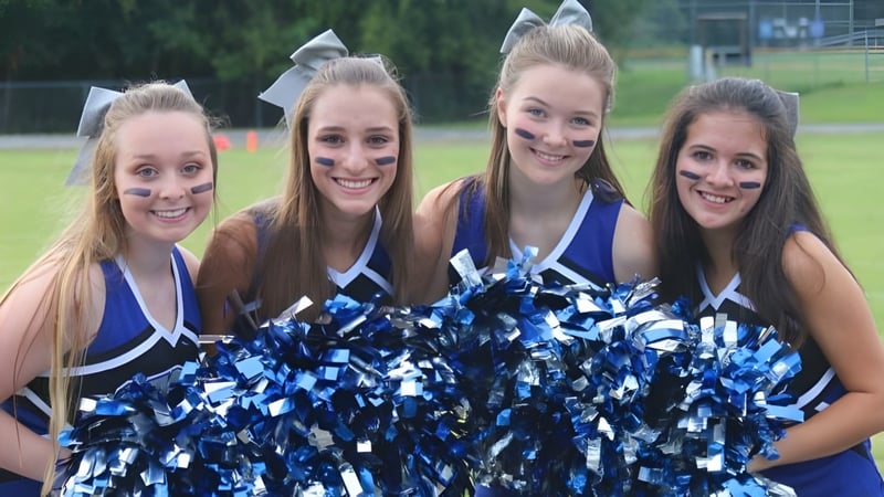 Vier Schülerinnen der Saint Francis Catholic Academy posieren im Freien mit blauen und weißen Pompons als Cheerleader.
