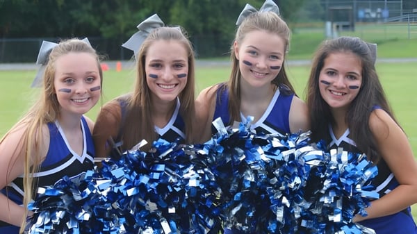 Vier Schülerinnen der Saint Francis Catholic Academy posieren im Freien mit blauen und weißen Pompons als Cheerleader.