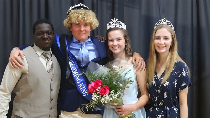 Eine Schülerin der Saint Francis Catholic Academy trägt eine Krone und hält einen Blumenstrauß vor einem dunklen Vorhang.