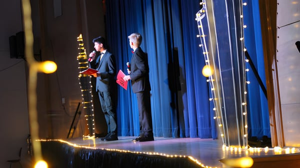 Zwei Personen in formeller Kleidung stehen auf der Bühne der Saint-Lambert International High School vor blauem Vorhang mit Lichterkette.