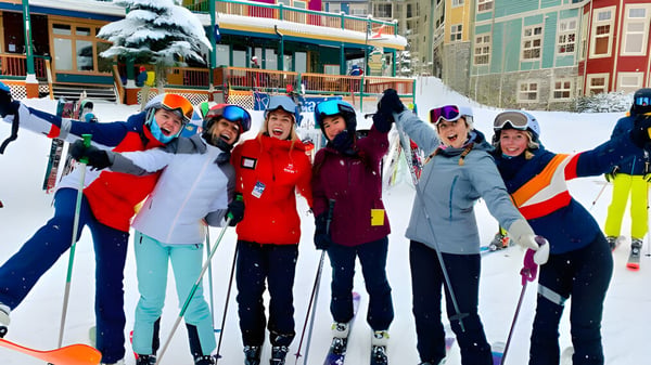Eine Gruppe von Schülerinnen und Schülern der Salmon Arm Secondary steht in bunter Skiausrüstung in einer verschneiten Landschaft vor Gebäuden und einem schneebedeckten Baum.