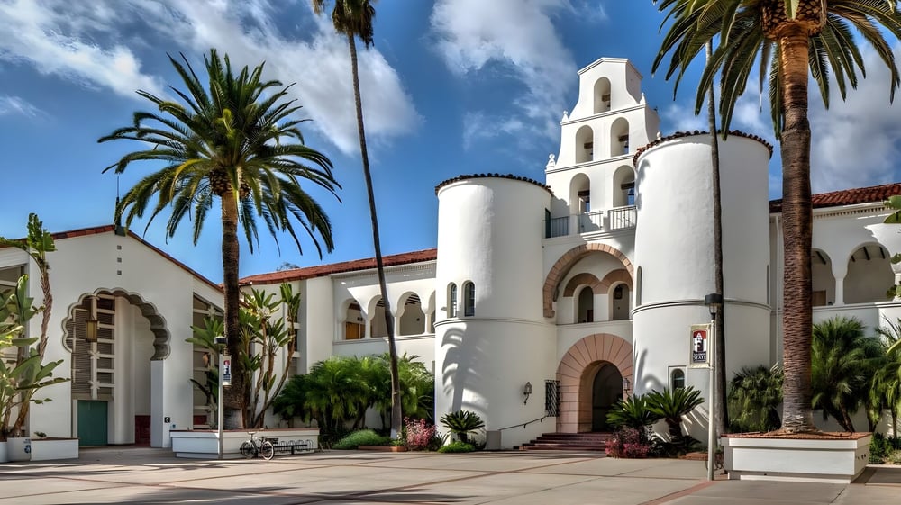 Das weiße Gebäude mit Glockenturm der San Diego State University ist von Palmen und Grün umgeben unter blauem Himmel.