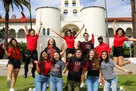 Eine Gruppe junger Studentinnen in roten Shirts posiert vor einem großen weißen Gebäude mit Glockenturm auf dem Campus der San Diego State University.
