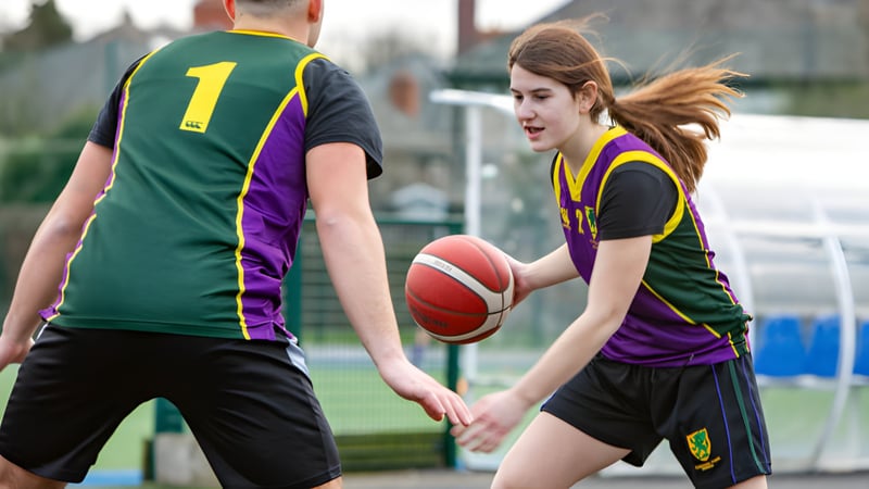 Zwei Schüler der Sandford Park School spielen Basketball auf einem Outdoor-Feld mit Zaun und Gebäuden im Hintergrund.
