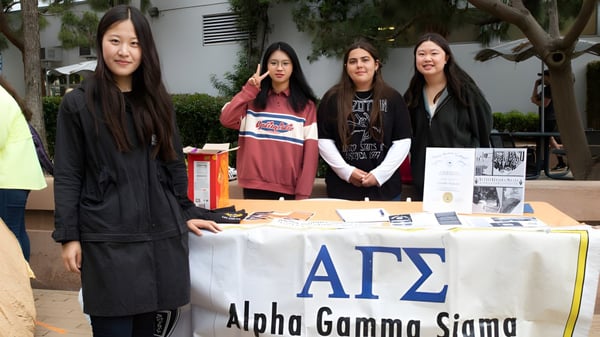 Vier Schülerinnen stehen hinter einem Tisch mit einem Alpha Gamma Sigma Banner auf dem Campus des Santa Monica College.