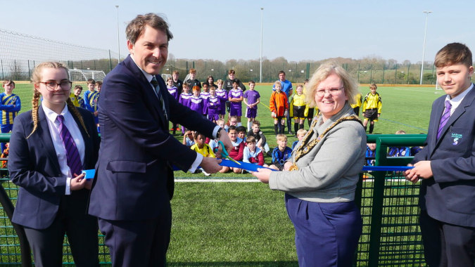 Eine Gruppe von Personen, darunter ein Mann im Anzug und eine Frau, stehen auf dem Fußballfeld der Sarum Academy, umgeben von Schülerinnen und Schülern in Sportuniformen.