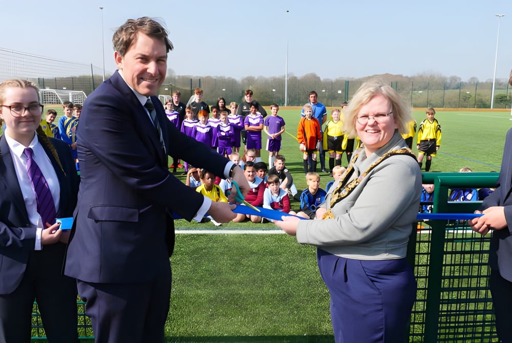Zwei Männer im Anzug geben sich auf dem Sportplatz der Sarum Academy vor einer Gruppe in bunten Uniformen die Hand.