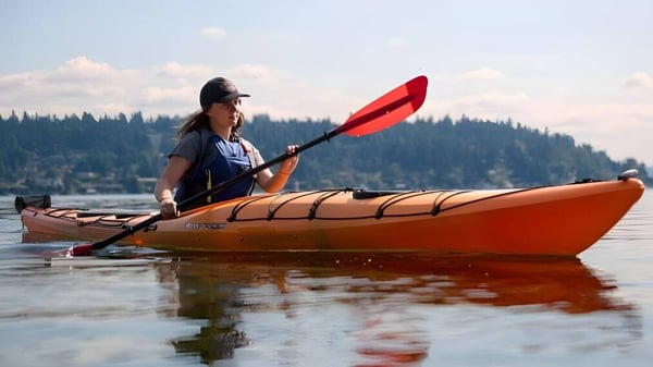 Eine Person paddelt in einem Kajak auf einem ruhigen See vor bewaldeten Bergen am Schull Community College.