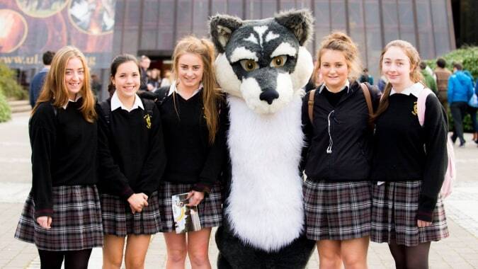 Eine Gruppe Schülerinnen posiert mit dem Wolf-Maskottchen vor einem Gebäude auf dem Campus der Scoil Aireagail.