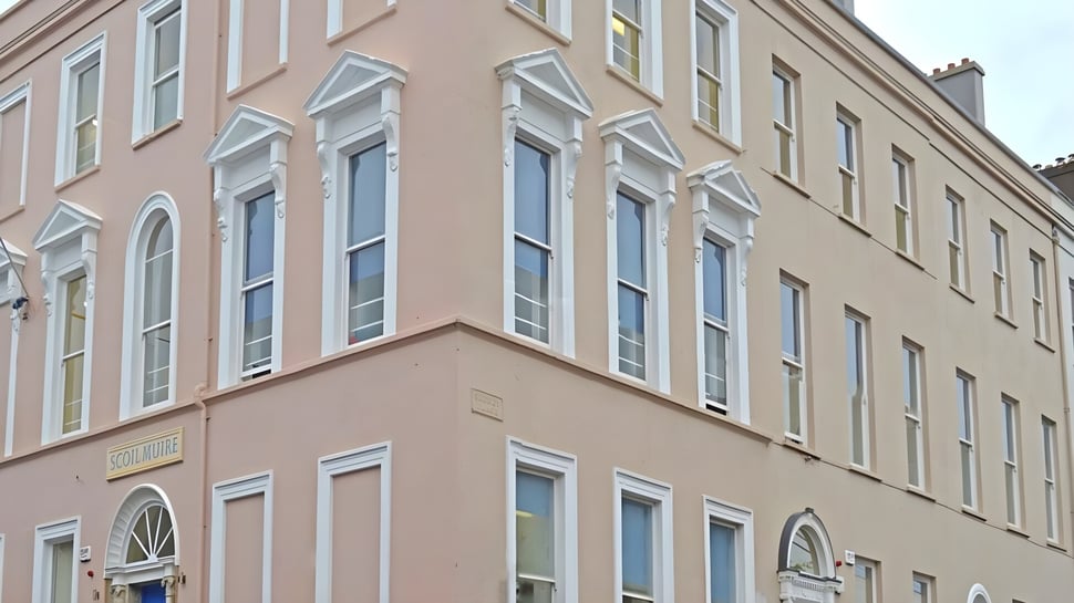 Das mehrstöckige Gebäude der Scoil Mhuire Cork mit großen Fenstern und rosa Fassade in urbaner Umgebung.