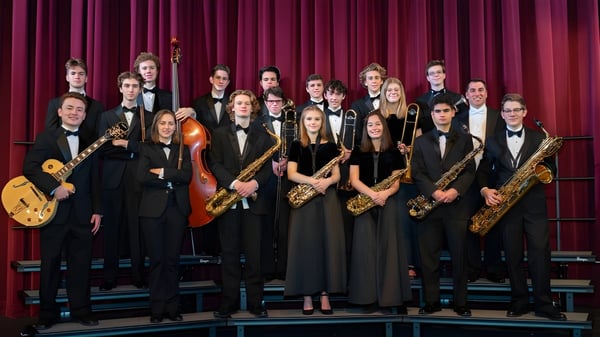 Eine Gruppe Musiker steht auf der Bühne mit roten Vorhängen und hält verschiedene Instrumente an der Scoil Mhuire gan Smál.