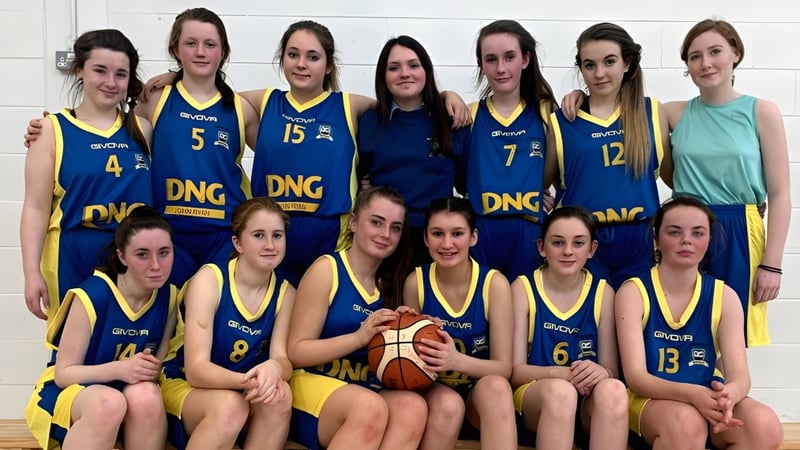 Eine Gruppe junger Schülerinnen der Scoil Mhuire Kanturk posiert als Basketballteam mit Ball vor einer schlichten Wand.