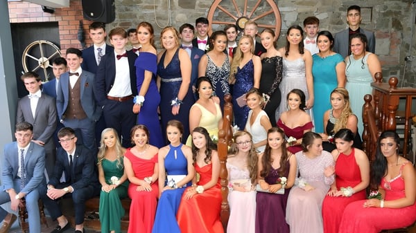 Eine Gruppe formell gekleideter Schülerinnen und Schüler auf dem Campus der Scoil Mhuire Kanturk vor rustikalem Hintergrund mit Holzrad.