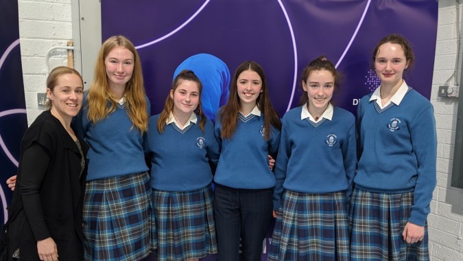 Eine Gruppe von Schülerinnen in blauen Schuluniformen steht vor einem violetten Hintergrund auf dem Campus der Scoil Mhuire (Trim).