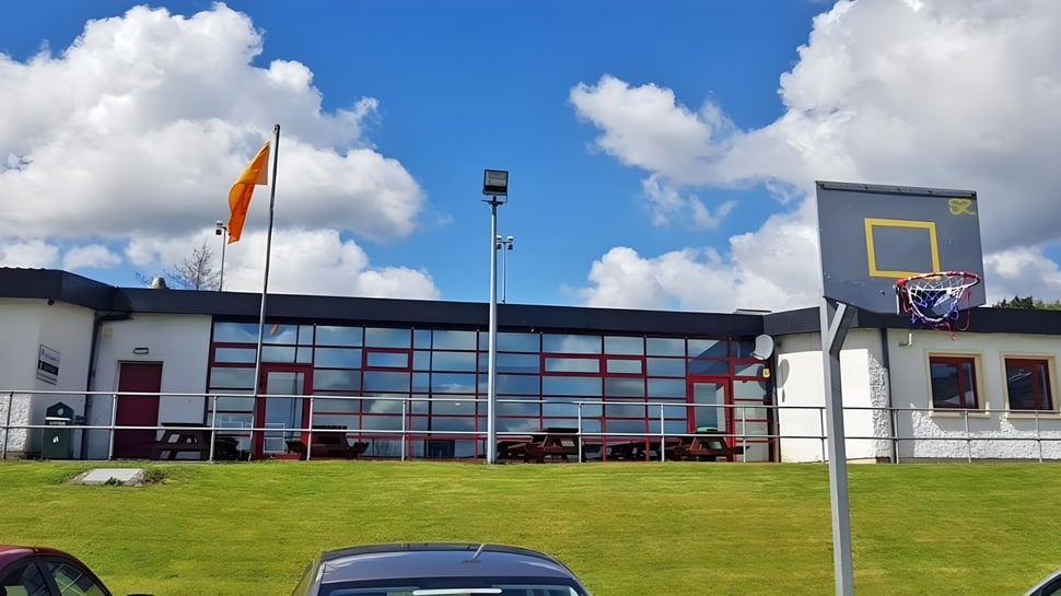 Das moderne Gebäude der Scoil Ruáin mit Basketballkorb steht auf einer grünen Wiese unter blauem Himmel.