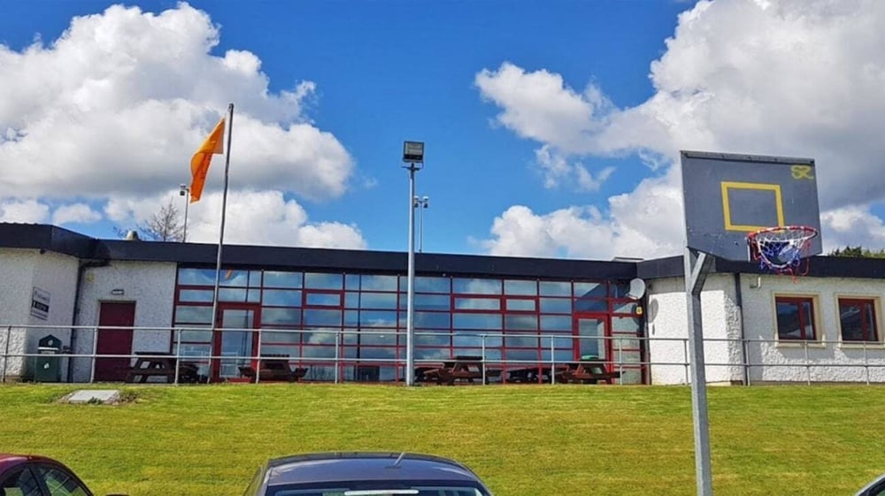 Das moderne Schulgebäude der Scoil Ruáin mit Basketballkorb steht auf einem Grasfeld unter einem bewölkten Himmel.