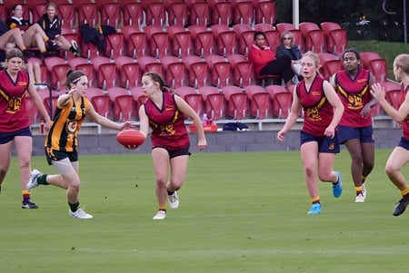 Eine Gruppe junger Athletinnen in roten und gelben Uniformen läuft auf einem Spielfeld beim Scotch Oakburn College vor Zuschauern.