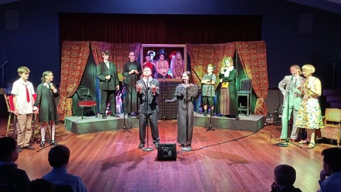 Auf der Bühne des Scots College stehen mehrere Schaufensterpuppen in bunten, vintage-inspirierten Outfits vor gemusterten Vorhängen und einem großen Bild an der Wand.