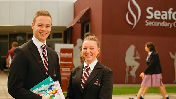 Zwei Schülerinnen und Schüler in Schuluniformen stehen vor dem Schild des Seaford Secondary College und halten ein Dokument.