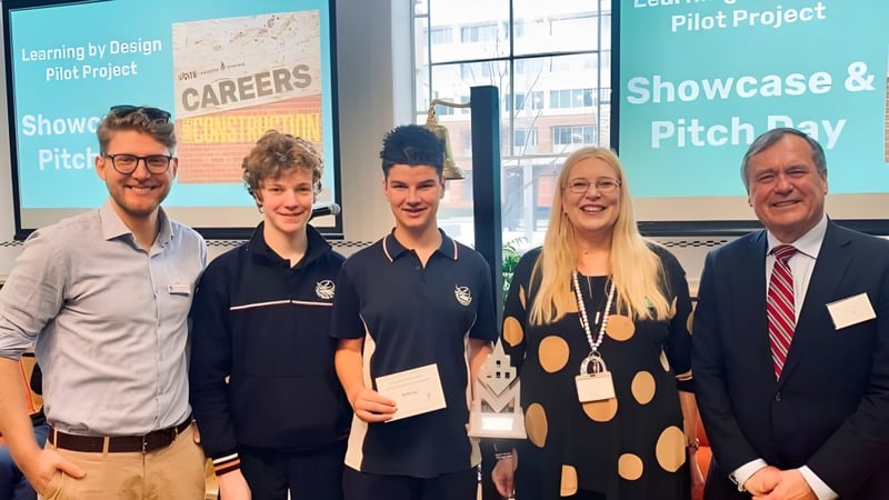 Eine Gruppe aus Schülern und einem Lehrer steht vor einem Informationsboard zum Learning by Design Pilot Project auf dem Campus der Seaton High School.